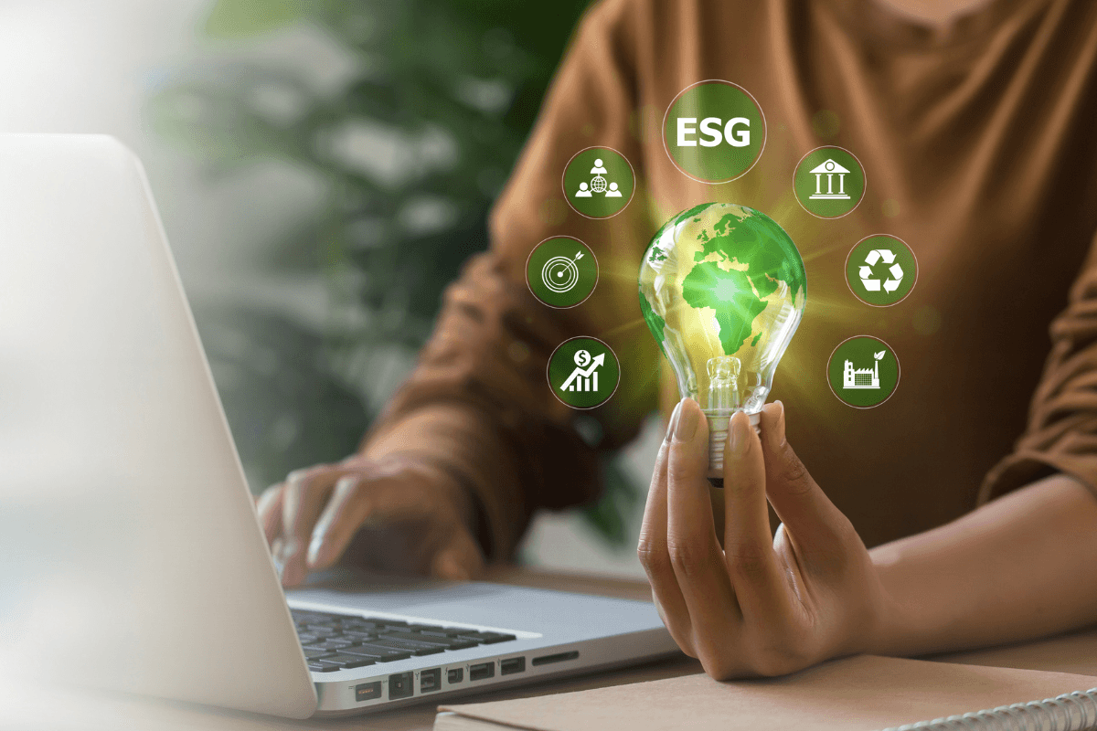 ESG Impact Criteria on Financing Woman typing with her right hand on a laptop and holding a light bulb reimagined as planet Earth (EMEA region is visible) with 7 bubbles shooting out of it (ESG at the top and other icons related to ESG, like recycling, factory, etc., around it) in her left hand.